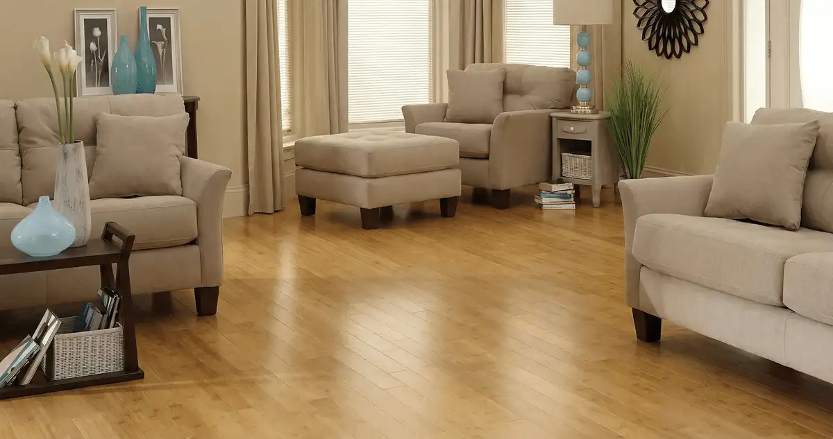 A living room with professionally installed bamboo flooring, beige sofas, a matching ottoman, a side table with a blue lamp, vases, framed pictures, and large windows with blinds and curtains in Richmond, VA